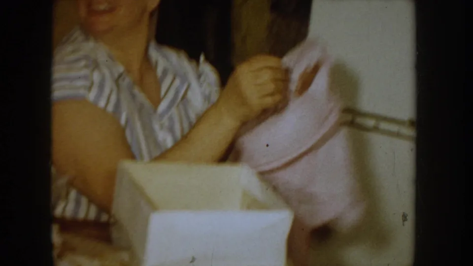 Close-up of a woman in a 1950s striped house dress lifting a pink clothing item from an open white gift box; Idaho, c.1955, 8mm.