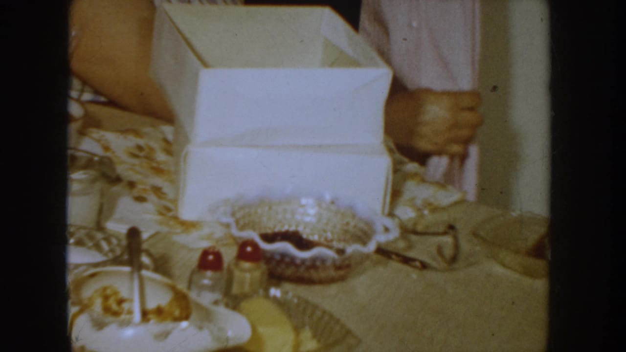 Mid-shot of a 1950s domestic table with an open white gift box, a decorative dish holding red items, condiment bottles, and a patterned tablecloth; second person partially visible at right.