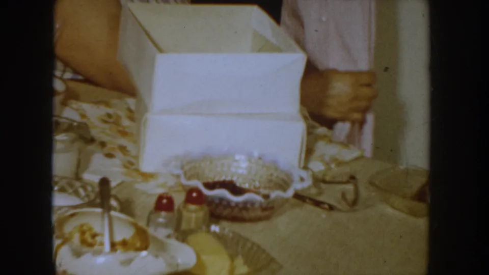 Mid-shot of a 1950s domestic table with an open white gift box, a decorative dish holding red items, condiment bottles, and a patterned tablecloth; second person partially visible at right.