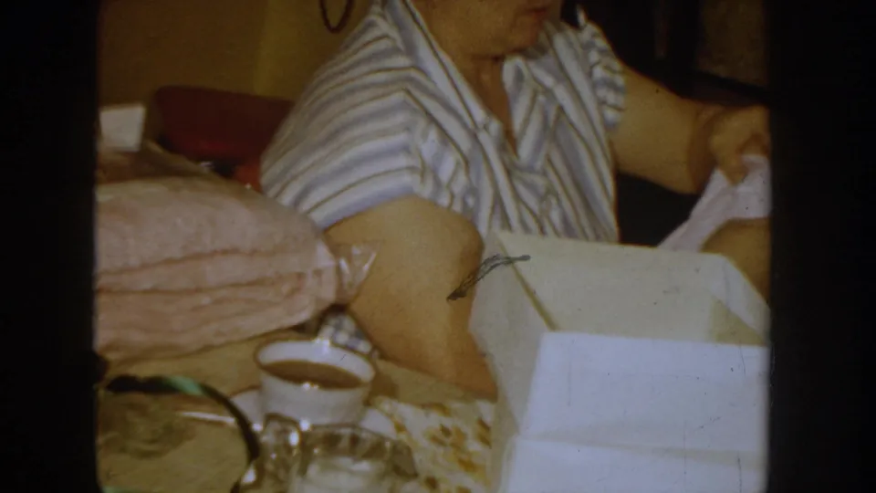 Side view of a woman in a striped 1950s house dress examining an open white gift box; pink fabric rests beside the box and a cup is visible at lower left.