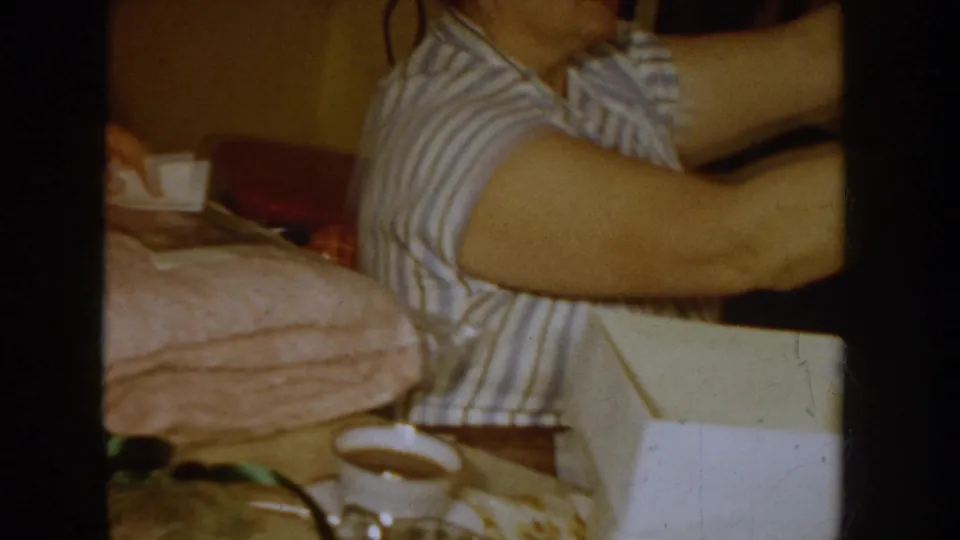 Woman in a striped 1950s house dress seated beside an open white gift box with pink garment fabric visible; cup at lower frame edge, private domestic interior, c.1955.
