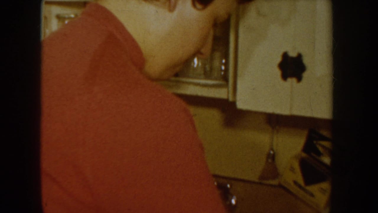 8mm home movie frame showing a figure in a red sweater beside a white period refrigerator in a kitchen interior, consistent with mid-1950s American domestic settings.
