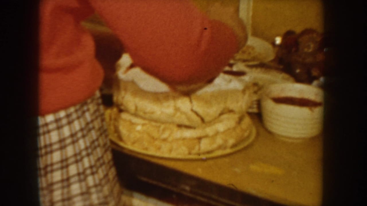 Person in red sweater and plaid skirt tends to a layered cream cake on a wooden table, with bowls visible in the background.