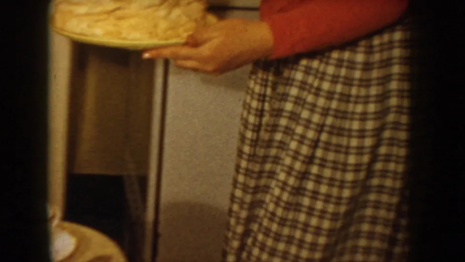 8mm home movie frame from circa 1955 Idaho showing a figure assembling a layer cake in a domestic kitchen, with a white period refrigerator visible behind them.