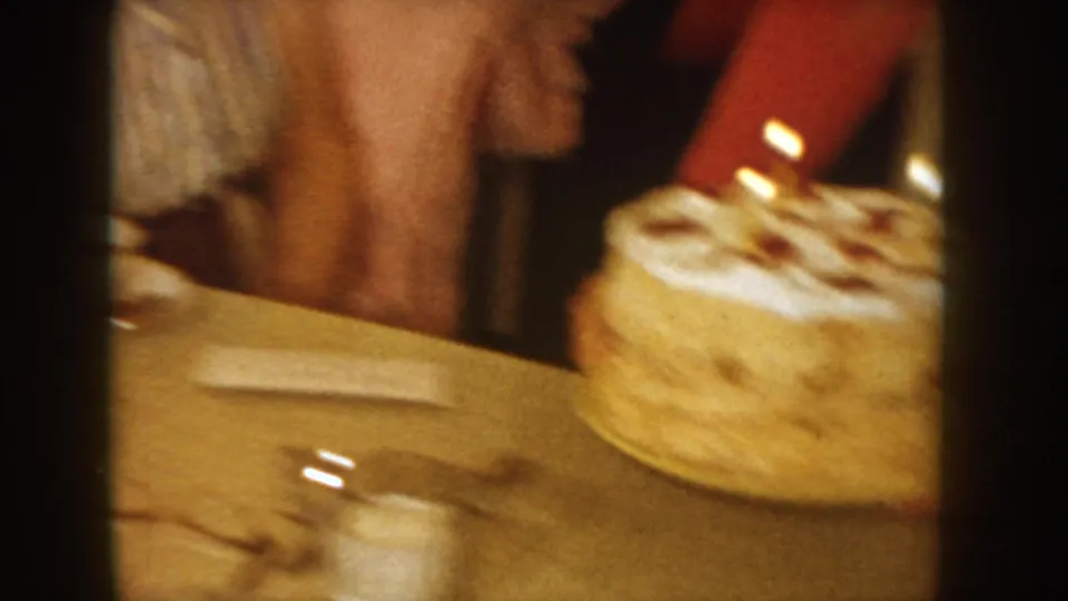 8mm home movie frame from circa 1955 Idaho showing a birthday cake with lit candles and red decorative accents on a table, with people gathered around in a domestic interior.