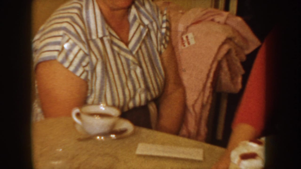 Two adults at a domestic table in what appears to be 1950s Idaho; the figure at left wears a blue-striped blouse and holds a white coffee cup; another in a red top is partially visible.