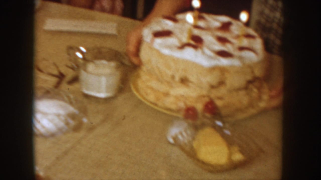 White-frosted layer cake with lit candles and red decorations on a plate, with a glass dish, fork, and condiment shakers on a light tablecloth.
