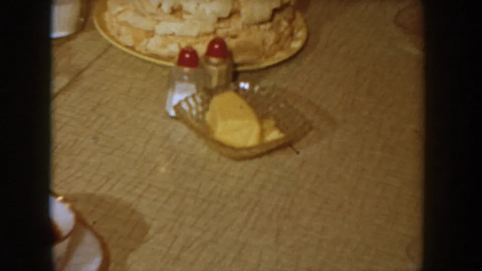 Table-level shot in warm-toned 8mm showing a white frosted cake, red-capped condiment shakers, and a butter dish, filmed at a 1955 Idaho domestic gathering.