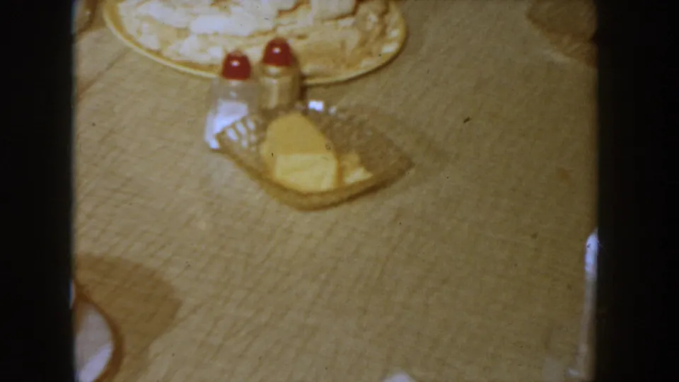 Continuing table-level 8mm shot of white frosted cake, red-capped salt and pepper shakers, and a butter dish on a light tablecloth at a 1955 Idaho domestic gathering.