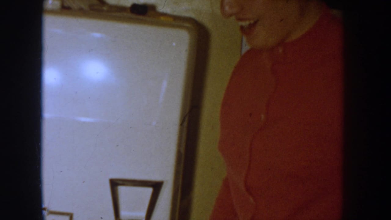 Smiling person in red button-up shirt near a white household appliance, Idaho home movie circa 1955, warm amber 8mm film tone.