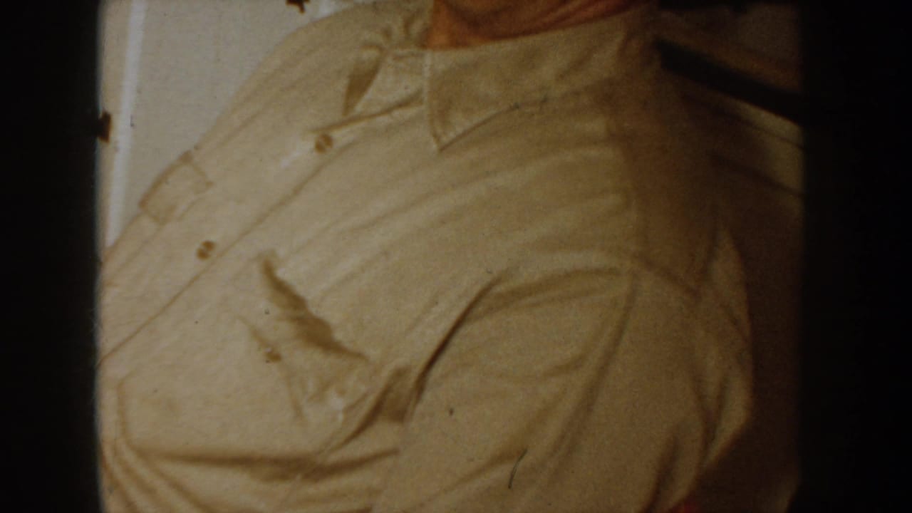 Close-up of a man's torso and collar in a light beige button-up shirt, heavy grain and warm color cast.