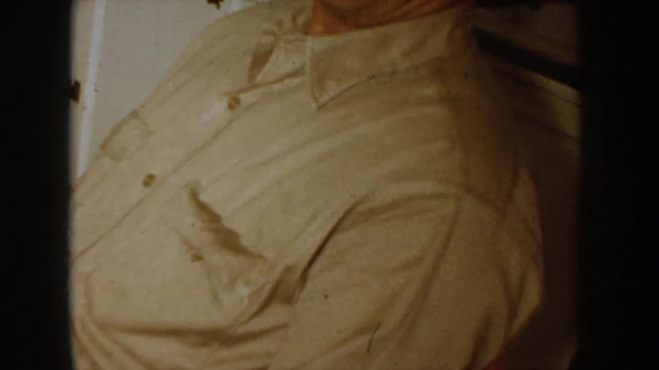 Tight mid-shot of a man's chest and collar in a light beige button-up shirt, Idaho home movie circa 1955, pronounced 8mm film grain.