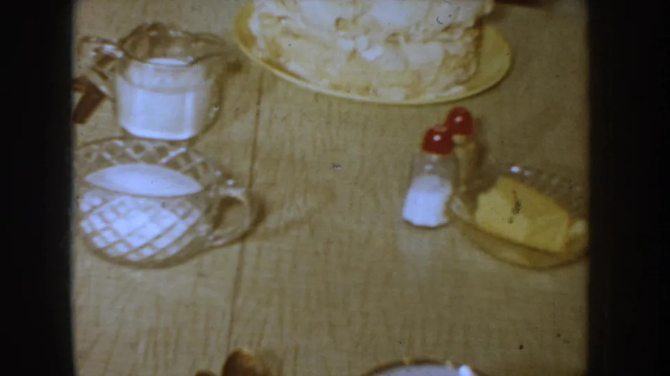 Close table-level view of a 1950s domestic table setting with glass pitcher, ceramic creamer, red-capped condiment shakers, butter dish, and layered frosted cake, Idaho circa 1955.