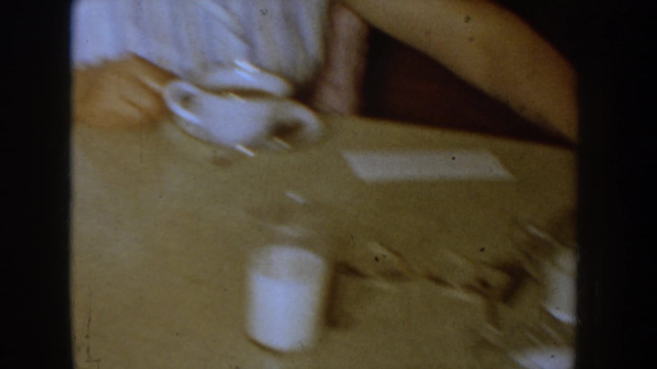 1955 Idaho 8mm home-movie frame showing a cropped adult at a table with a cup, paper, and warm interior lighting.
