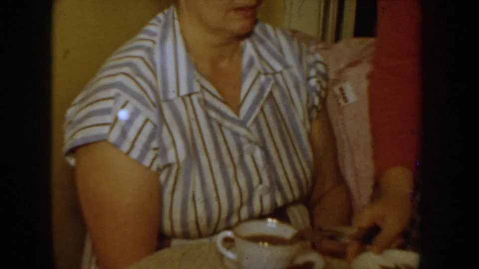 1955 Idaho home-movie frame showing a woman in a striped blouse seated at a table with a teacup while another adult is partly visible beside her.