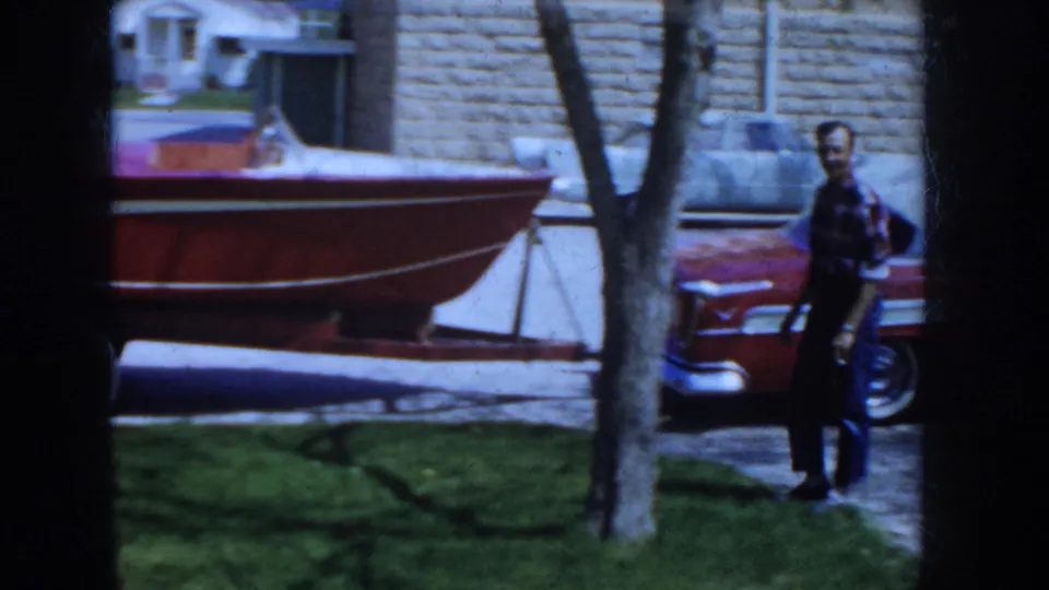 1955 Idaho 8mm frame showing a man outdoors near a classic red car and a boat on a trailer beside a building wall, trees, and grass.