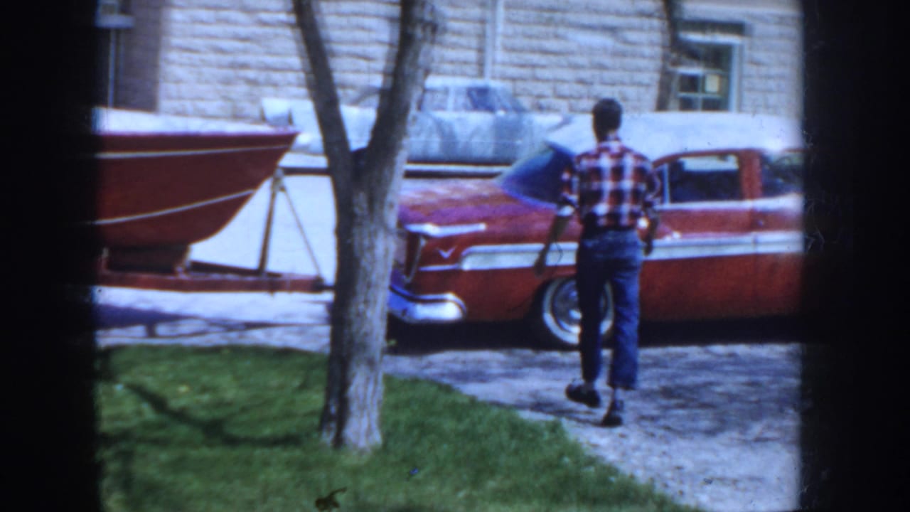 Man in plaid shirt and blue jeans approaches a two-tone red and white mid-1950s American sedan on a gravel driveway, red boat on trailer at left, cut-stone building behind; Idaho, c.1955.
