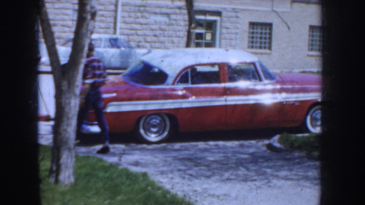 Man in plaid shirt continues past the rear of a two-tone red and white 1950s sedan; stone building and bare trees visible in background.