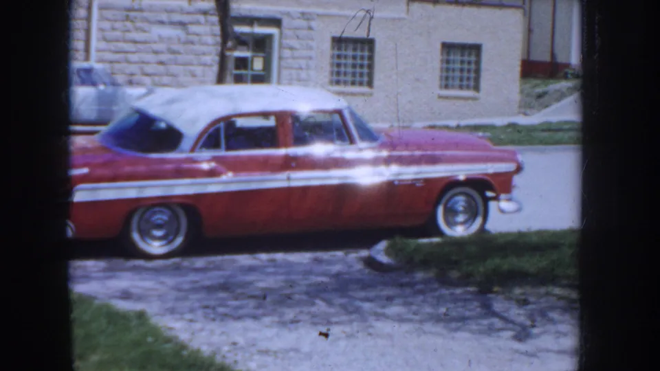 Side profile of a two-tone red and white mid-1950s American sedan with whitewall tires and chrome side trim, parked on a gravel driveway in front of a cut-stone masonry building; Idaho, c.1955.