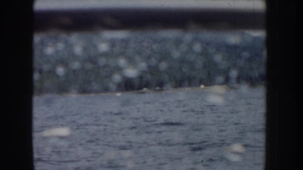 8mm home movie frame from a moving boat on an Idaho lake, circa 1955; dark window borders frame a view of rippled water and a distant blurred treeline.