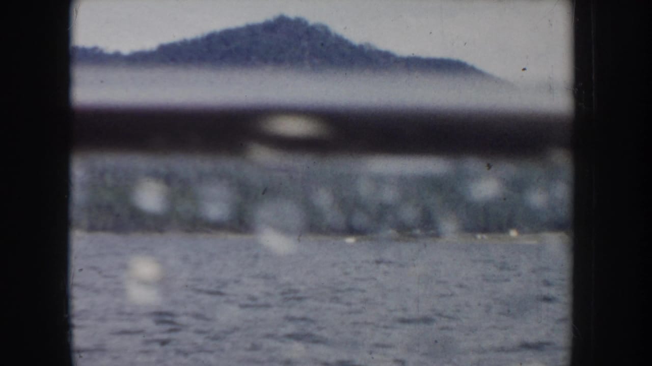 Boat window view with a rounded forested hill against a pale sky, lake water in the foreground, and a horizontal boat railing visible at the top edge.