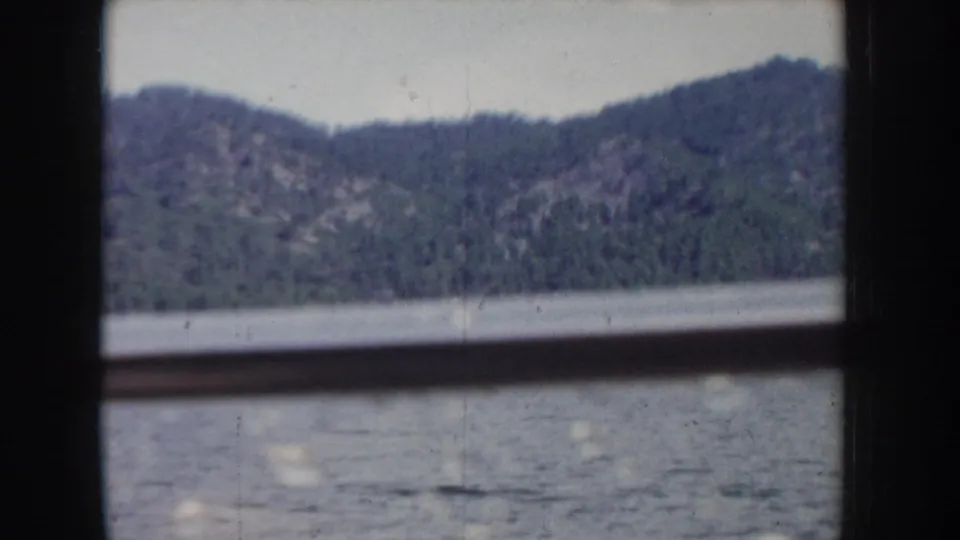 8mm home movie frame through a boat window, circa 1955 Idaho; steep conifer-covered hillsides with exposed rocky faces rise above calm lake water.
