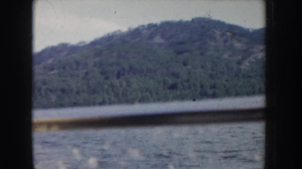 8mm home movie frame through a boat window, circa 1955 Idaho; forested hills above calm water with a possible shoreline structure at the lower right edge.