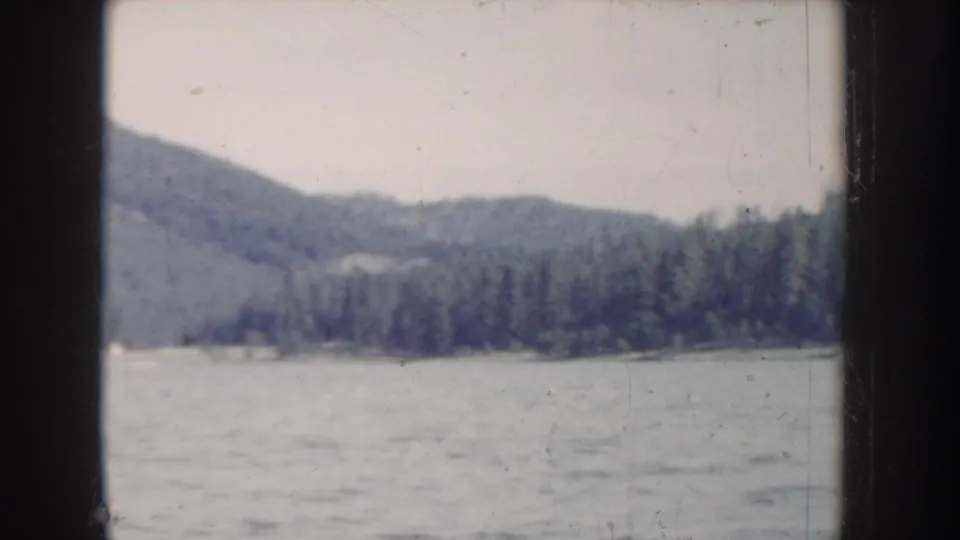 1950s 8mm home movie frame showing a rounded mountain with conifer tree line and calm lake in what is described as Idaho, USA; notably desaturated color.