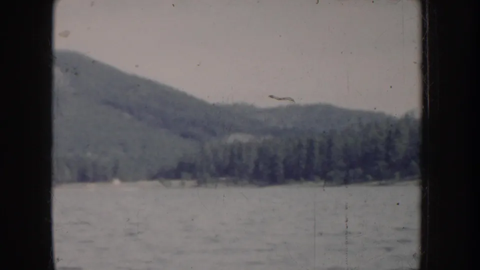 1950s 8mm home movie frame of a conifer-covered mountain and lake in what is described as Idaho, USA; pan continues to reveal wider mountain silhouette.
