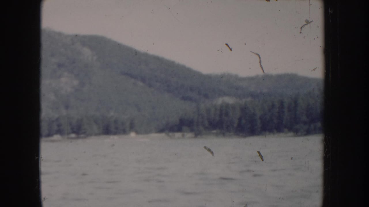 8mm home-movie circa 1955, Idaho — boat-level view of choppy mountain lake with dense conifer hillside under overcast sky.