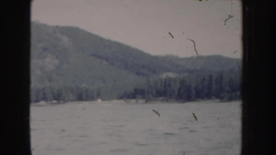 8mm home-movie circa 1955, Idaho — boat-level view of choppy mountain lake with dense conifer hillside under overcast sky.