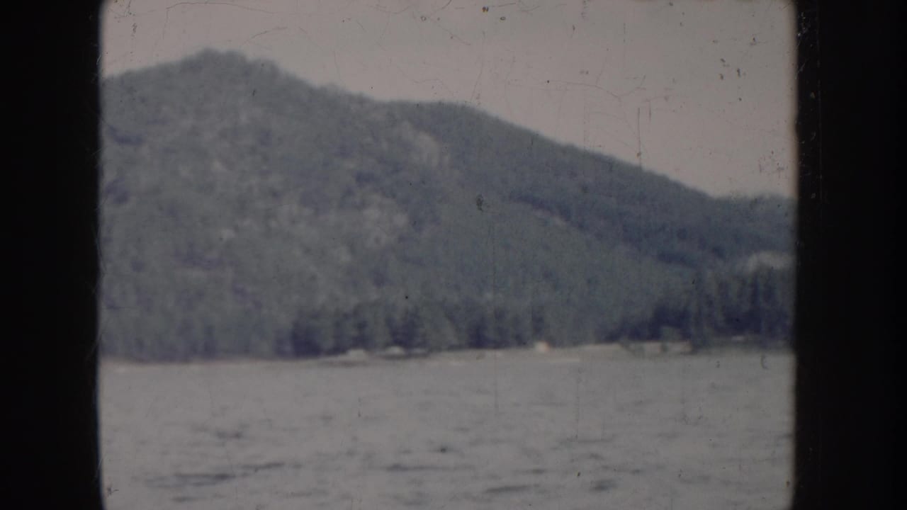 Boat-level view of choppy lake with forested mountain and faint structure at treeline base, under overcast sky.