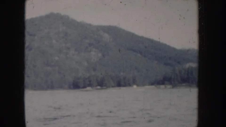 8mm home movie circa 1955, Idaho — lake surface and forested mountain from moving boat; faint structure visible at the shoreline base.