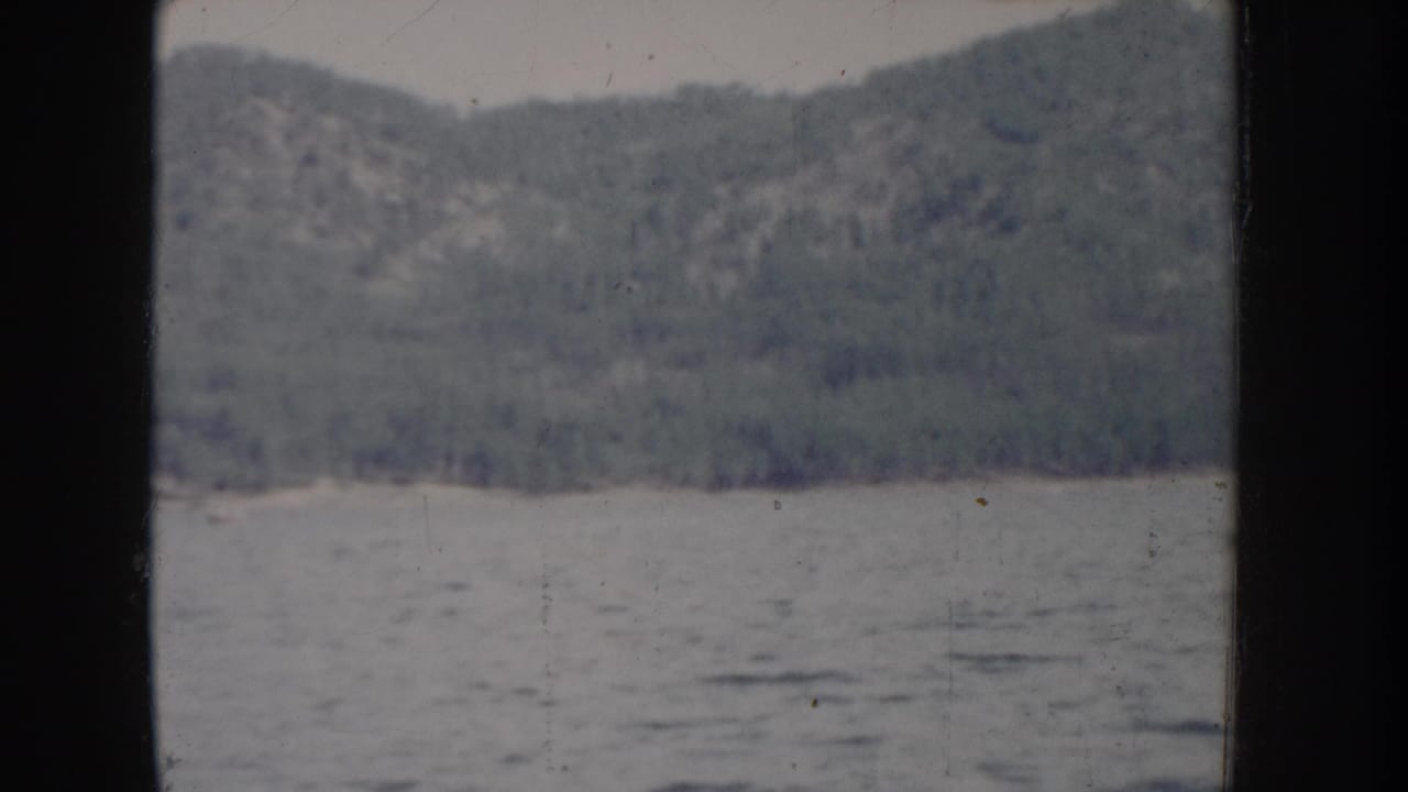 Opening 8mm frame of a calm Idaho lake circa 1955, with a dense conifer-covered hillside rising above the water and heavy blue-gray color fading throughout.