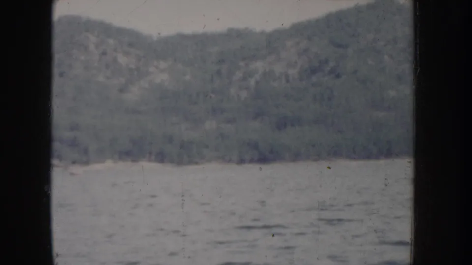 Opening 8mm frame of a calm Idaho lake circa 1955, with a dense conifer-covered hillside rising above the water and heavy blue-gray color fading throughout.
