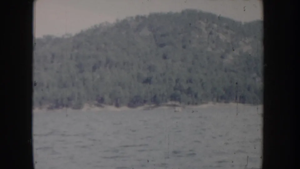 Mid-clip 8mm frame of Idaho lake circa 1955; the forested hillside occupies a greater portion of the frame, with pronounced grain and consistent blue-gray fading.