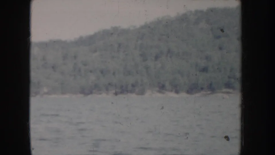 Closing 8mm frame of Idaho lake footage circa 1955, showing forested hillside above calm water with dark vignette borders and heavy color fading consistent throughout the clip.