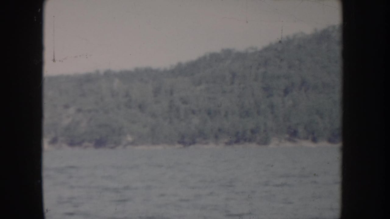 POV river shot from a 1955 Idaho home movie showing calm water and a tree-covered hillside in faded 8mm color.