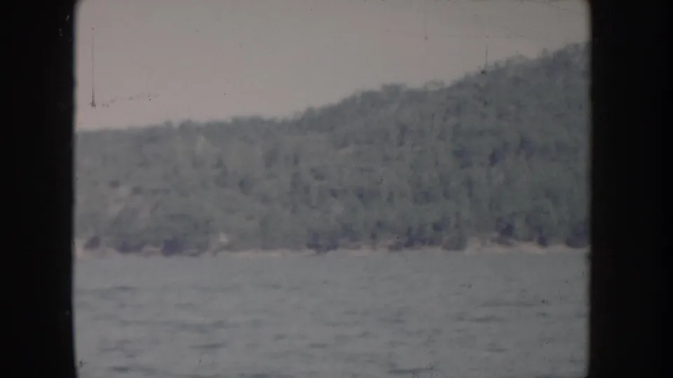 POV river shot from a 1955 Idaho home movie showing calm water and a tree-covered hillside in faded 8mm color.