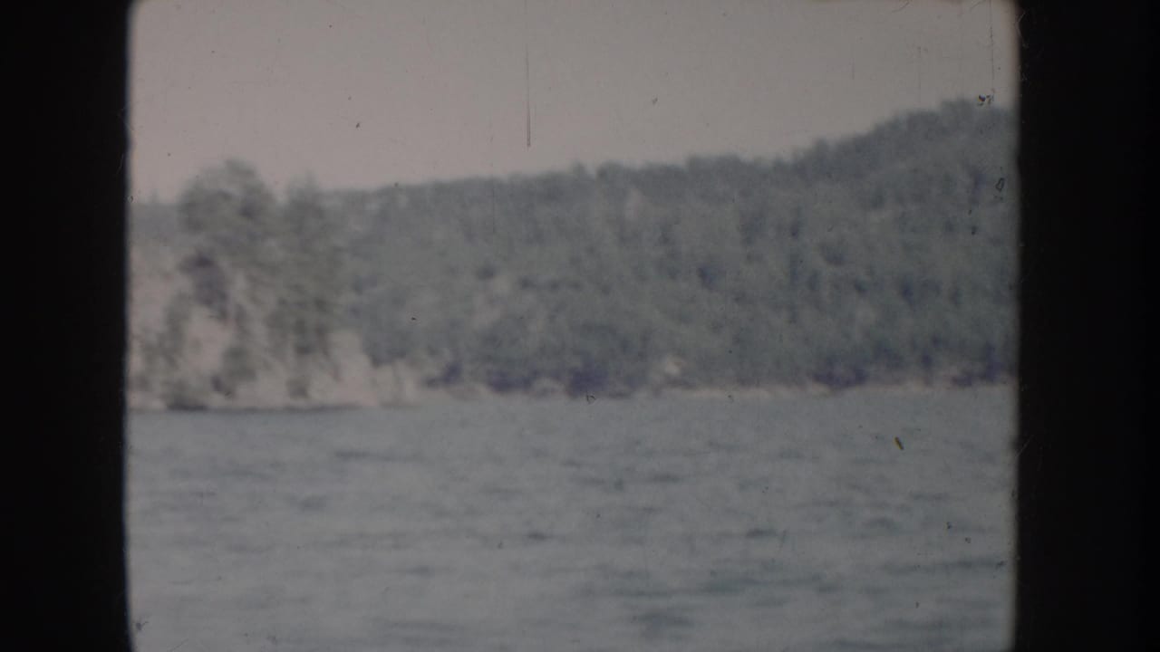 Calm river with wooded hills and a pale rocky outcrop on the far bank, 8mm color film.
