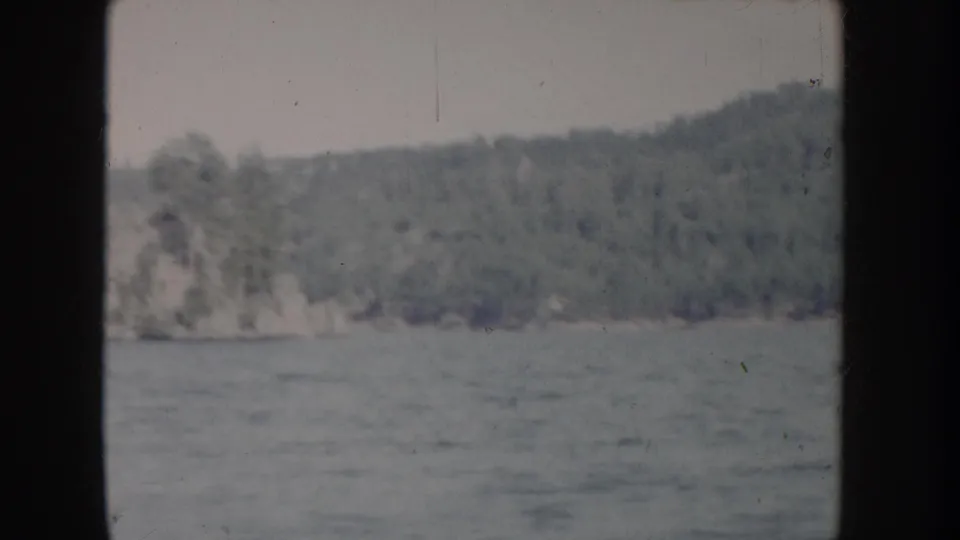 Moving-boat POV of a wooded river bank with a rocky cliff outcrop, Idaho waterway circa 1955 in faded 8mm color.