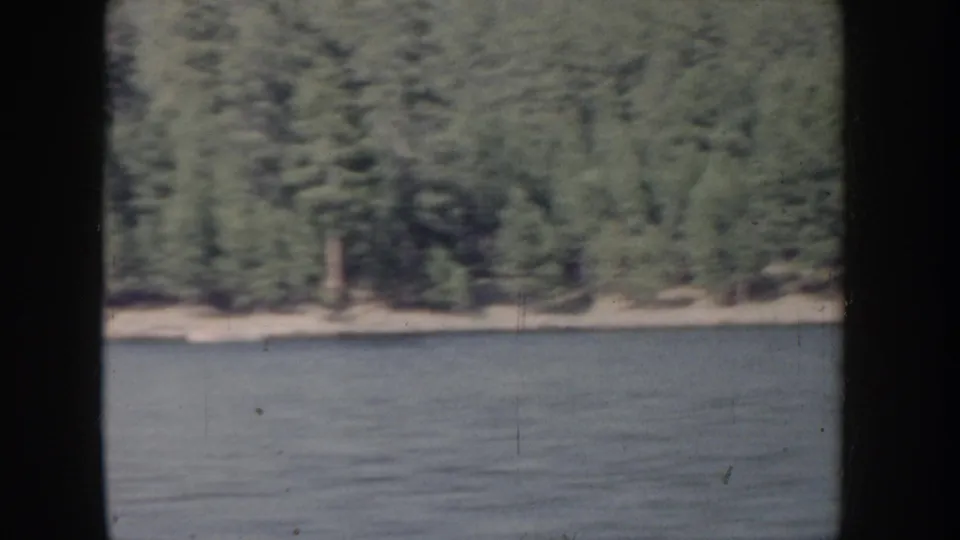 Mid-clip frame from Idaho lake footage circa 1955; calm water foreground with a dense evergreen tree line and sandy shore under muted 8mm color.