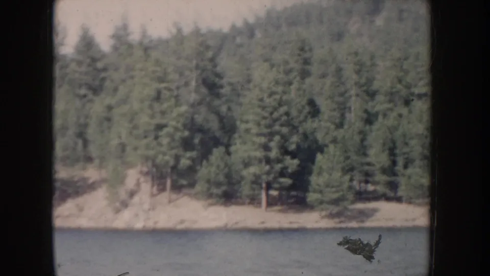 Final frame of Idaho lake clip circa 1955; calm water, dense conifer shoreline, sandy bank, and a small unidentified dark shape in the lower-right corner.