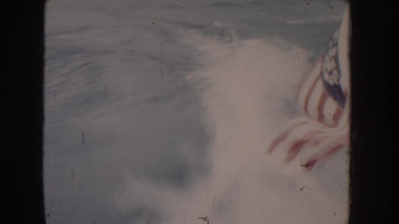 American flag close-up from a 1955 Idaho home movie; red and white stripes and blue star canton visible on heavily faded 8mm stock.