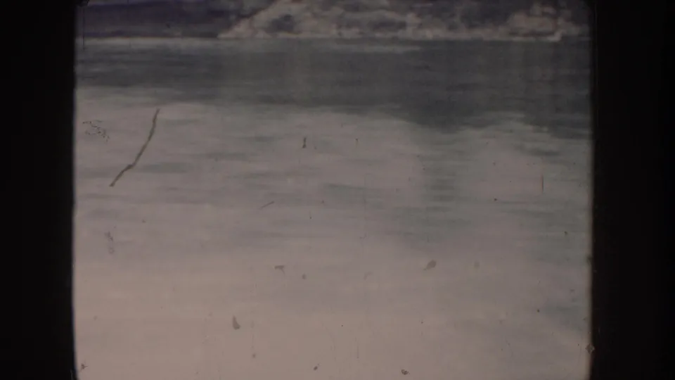 Lake surface filmed from a moving boat in 1955 Idaho; pale pink-beige color fading typical of aged 8mm film, with distant shore visible at top of frame.