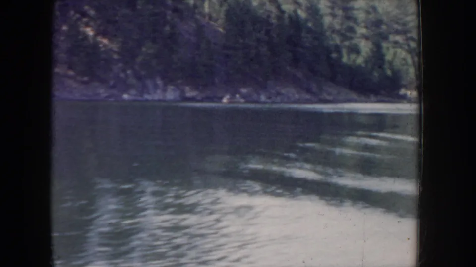 View from a moving boat of a forested river canyon, circa 1955 Idaho; evergreen-covered hillside with rocky base, boat wake in foreground, blue-purple aged 8mm color cast.