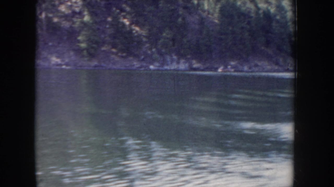8mm home movie from Idaho circa 1955 — calm lake water in the foreground with a dense evergreen hillside on the far shore, shot from a moving boat.
