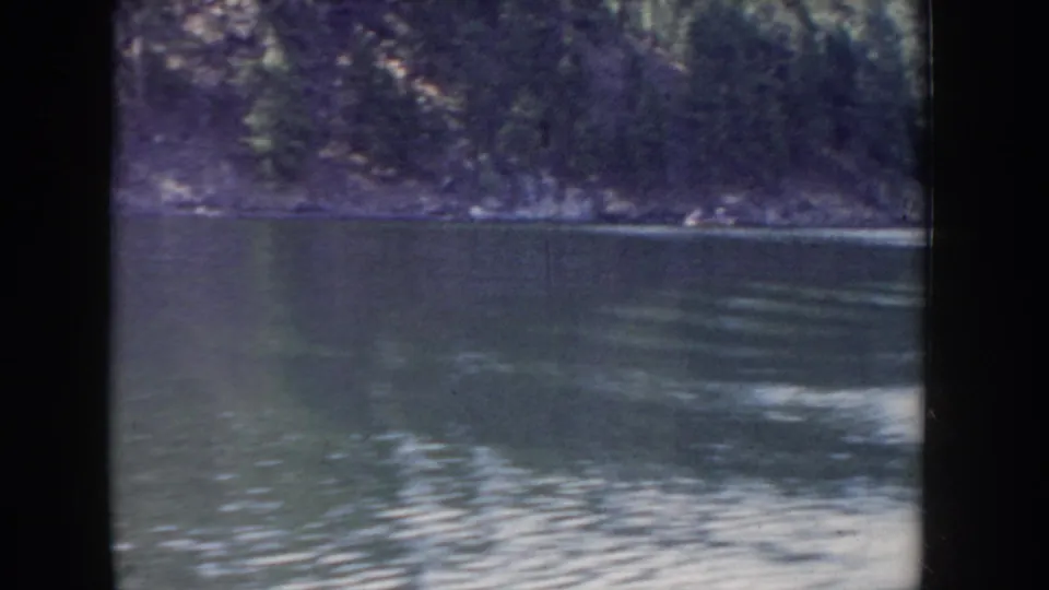 8mm home movie from Idaho circa 1955 — calm lake water in the foreground with a dense evergreen hillside on the far shore, shot from a moving boat.