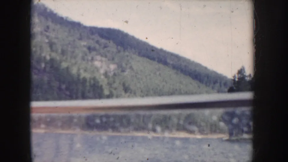 8mm transition frame from what metadata describes as Idaho, 1955 — a steep evergreen hillside alongside water with an overcast pale sky, likely filmed from a boat or road.