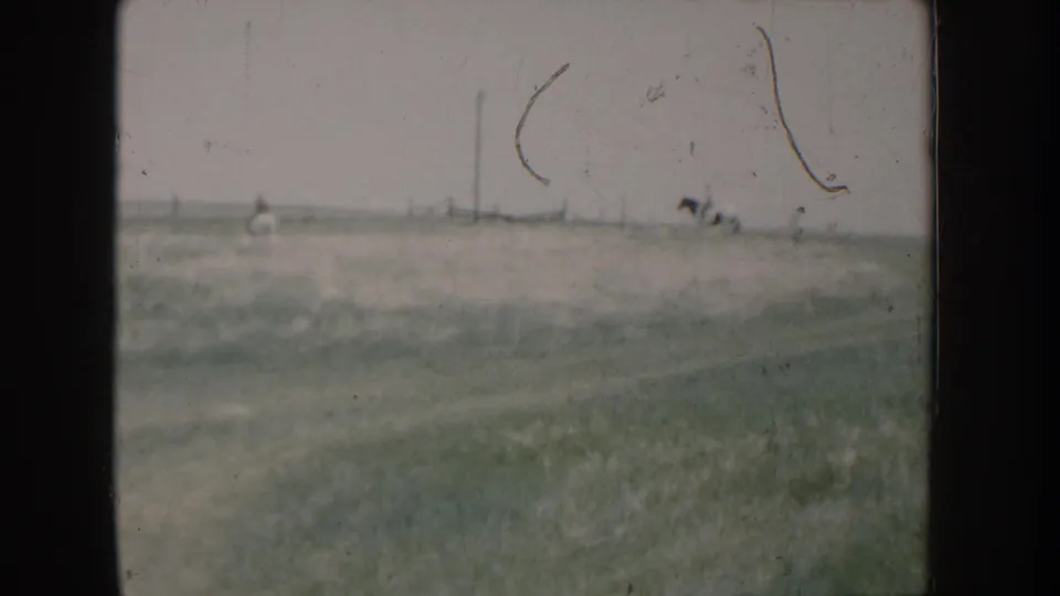 8mm home movie frame showing open rural grassland in Idaho circa 1955; a small distant figure may be a person on horseback, with utility poles and prominent film-hair scratches visible.